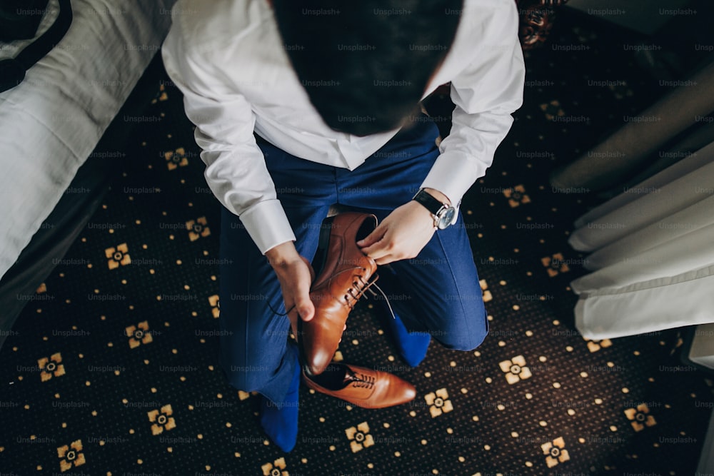 Stylish groom in blue suit putting on brown shoes near window in hotel room, top view. Morning preparation before wedding ceremony. Man getting ready before luxury event