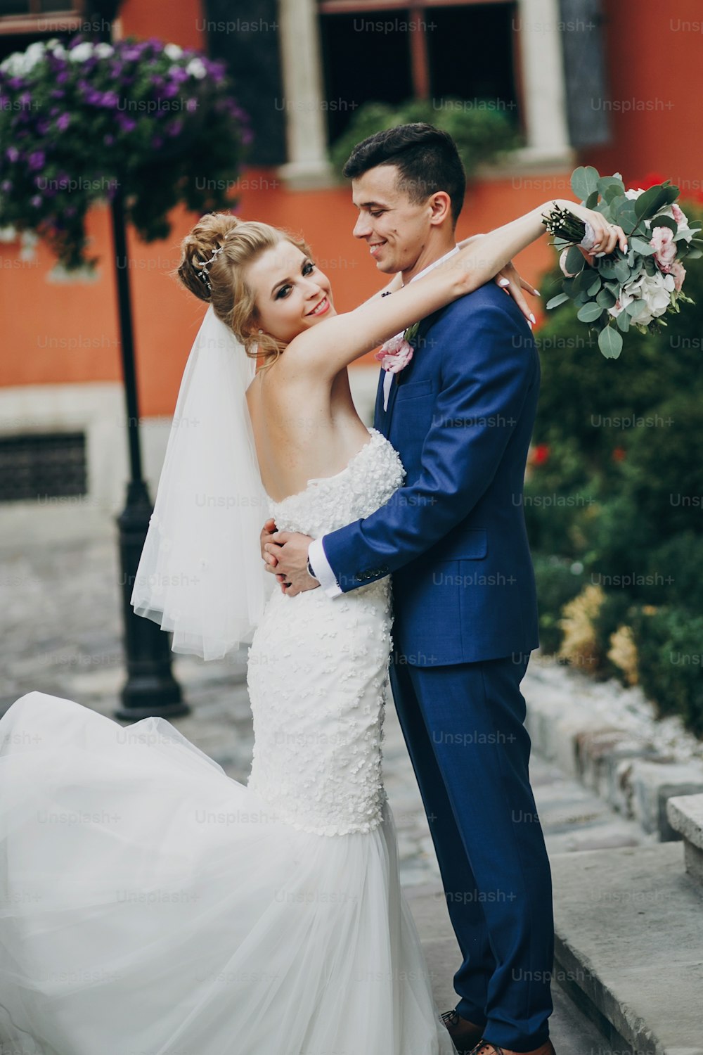 Noiva e noivo elegantes posando e sorrindo na ensolarada rua da cidade europeia. Lindo casal de recém-casados se abraçando em prédios antigos. Momento romântico