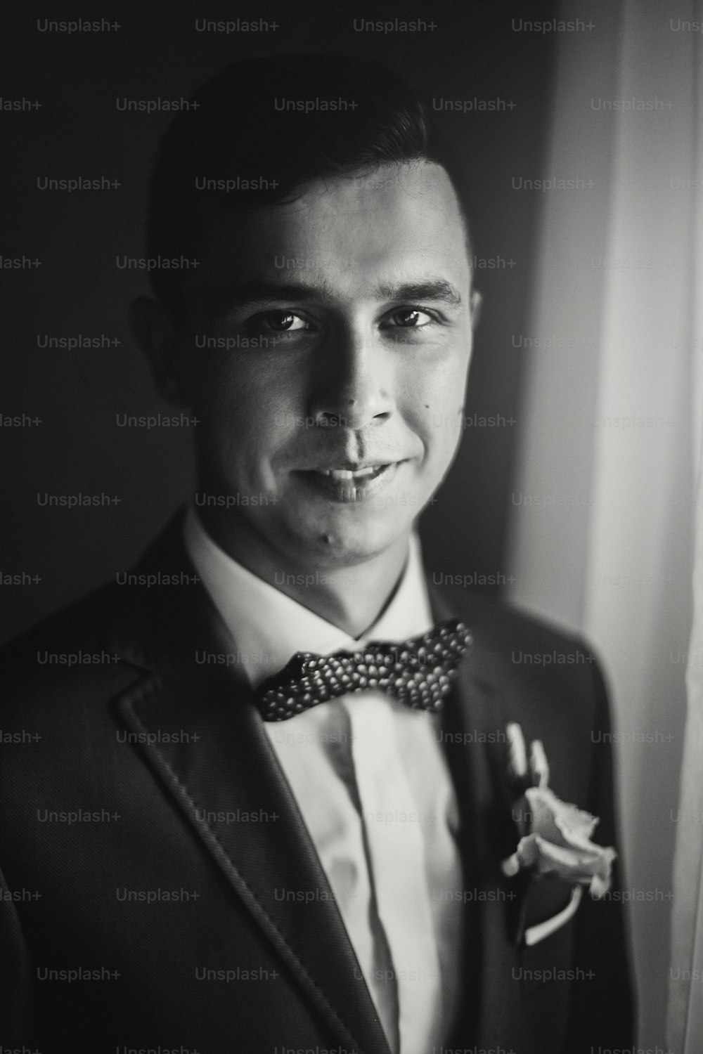 Stylish happy groom in blue suit, with bow tie and boutonniere posing and smiling near window in hotel room. Morning preparation before wedding ceremony