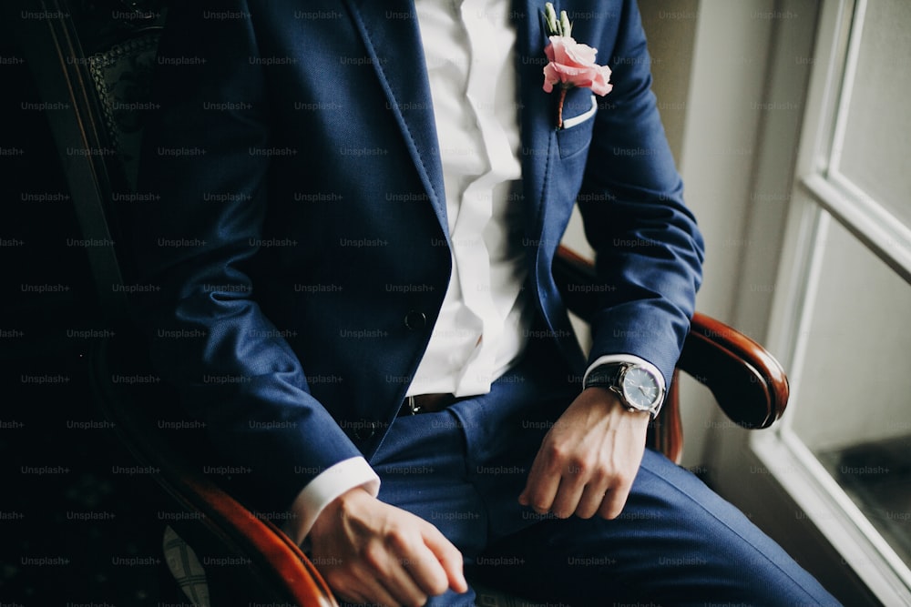 Noivo elegante em terno azul, com gravata borboleta e boutonniere com rosa rosa sentado na cadeira perto da janela no quarto de hotel. Preparação matinal antes da cerimônia de casamento
