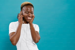 Stylish, cheerful black woman with pleasure listening music with closed eyes with headphones. Young afro woman in white t shirt posing on blue background with copy space.