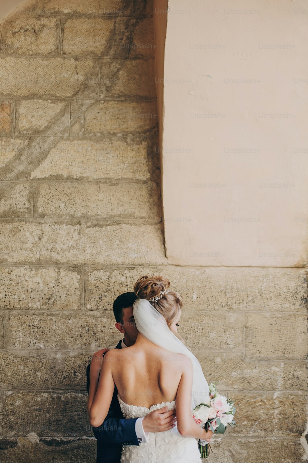 Stylish groom gently hugging beautiful bride back in city street. Gorgeous wedding couple of newlyweds sensually embracing at old buildings, back view. Romantic moment