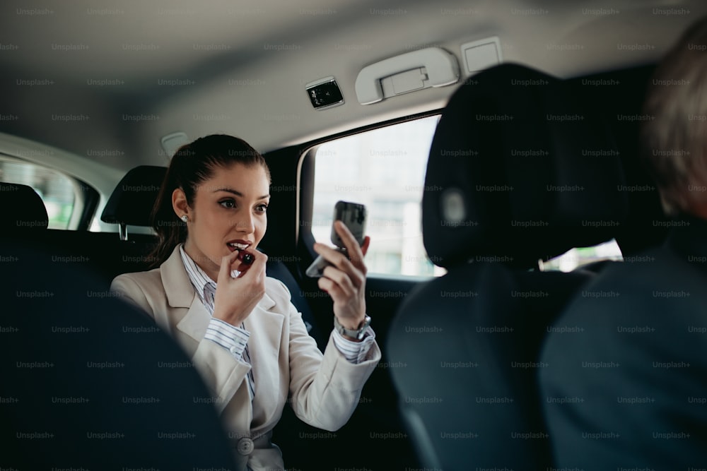 Gut aussehende junge Geschäftsfrau, die auf dem Rücksitz in einem Luxusauto sitzt und ihr Make-up repariert. Transport im Unternehmenskonzept.