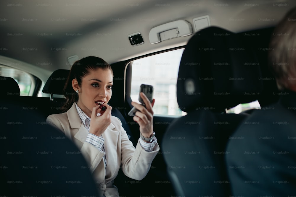 Good looking young business woman sitting on backseat in luxury car and fixing her makeup. Transportation in corporate business concept.