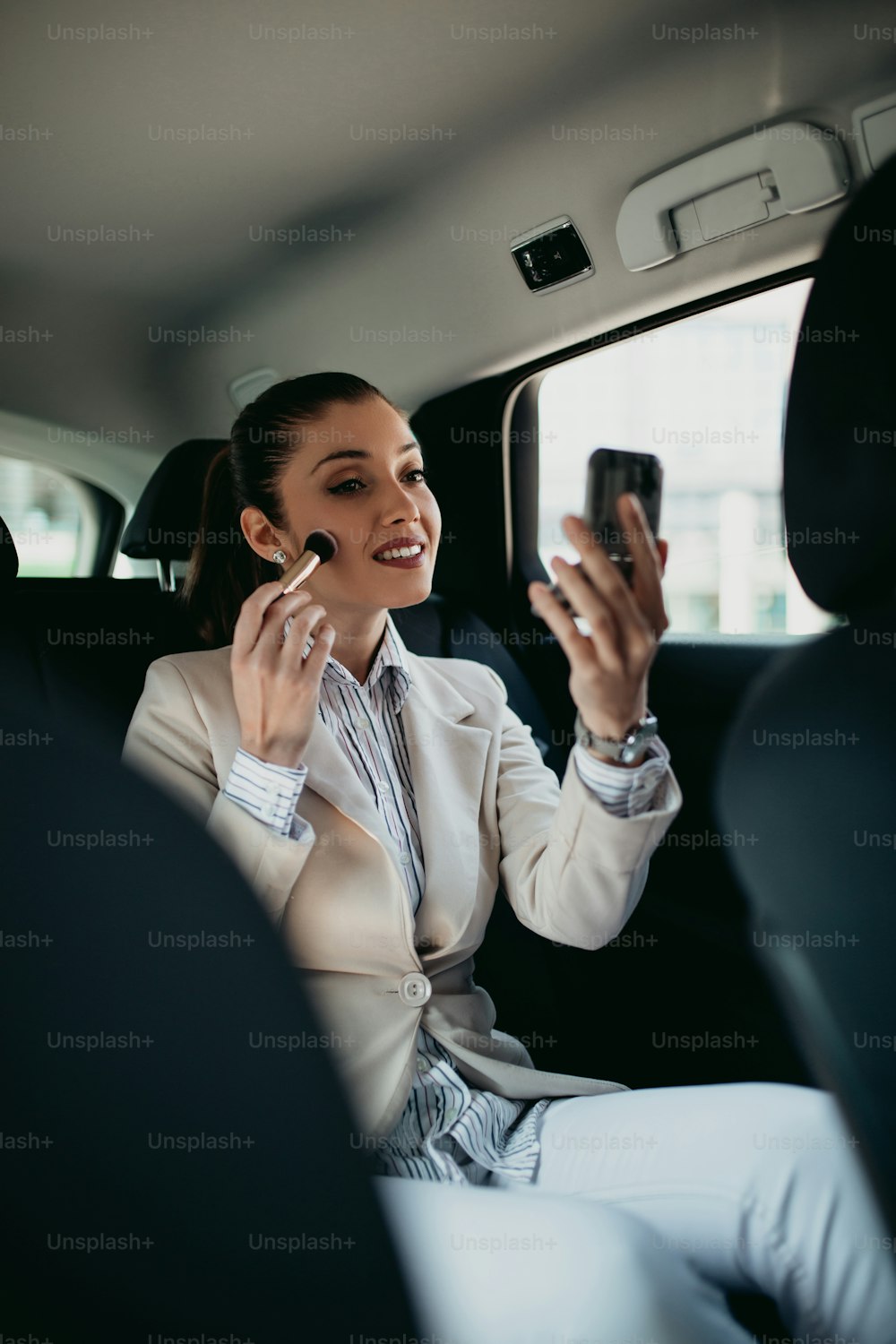 Gut aussehende junge Geschäftsfrau, die auf dem Rücksitz in einem Luxusauto sitzt und ihr Make-up repariert. Transport im Unternehmenskonzept.