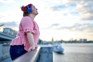Young fat woman enjoying music and standing on bridge in the city near river