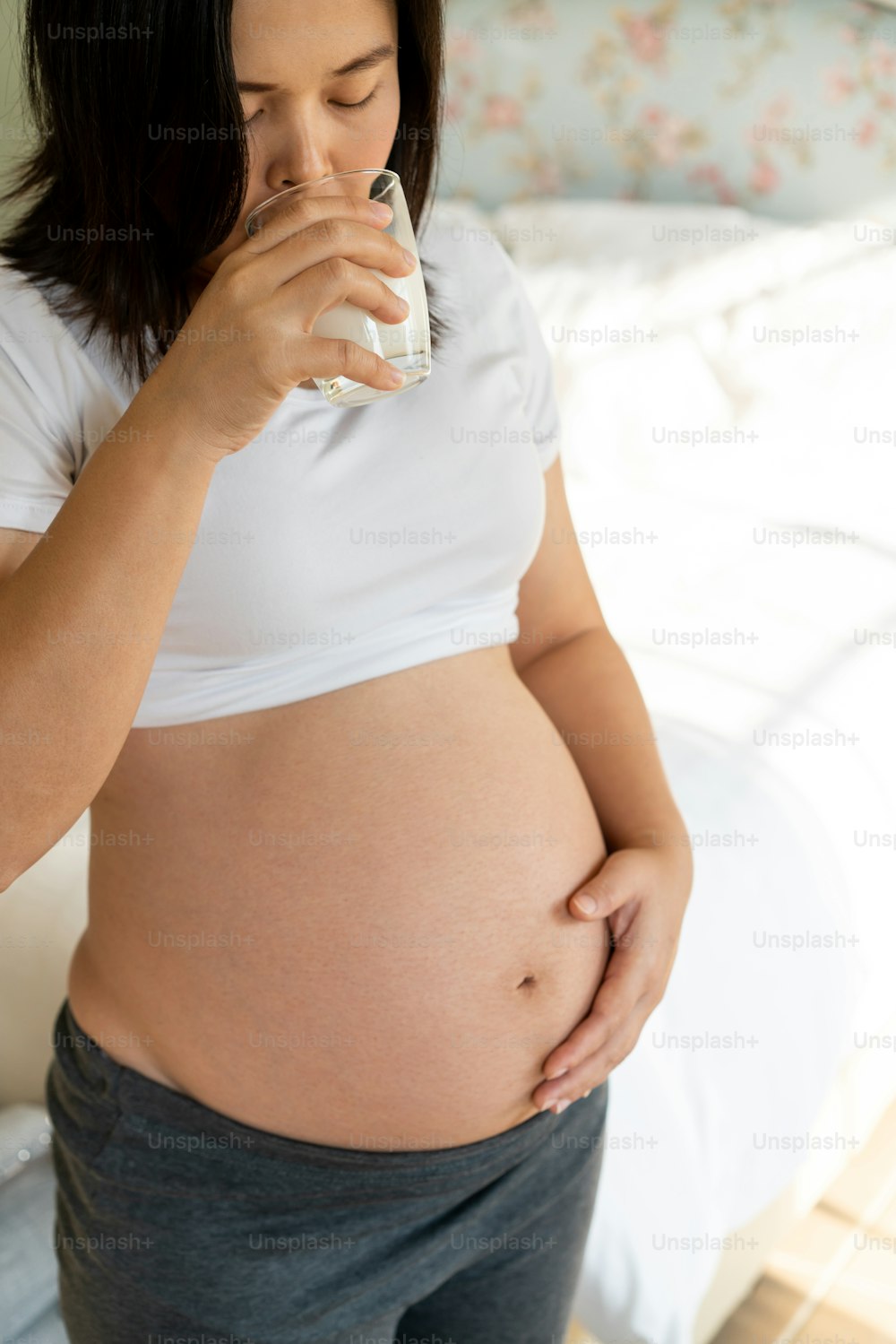 Happy pregnant woman drinks milk in glass at home while taking care of her child. The young expecting mother holding baby in pregnant belly. Calcium food nutrition for strong bones of pregnancy.