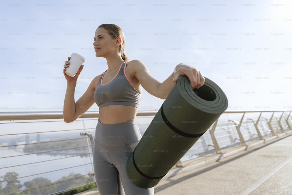 Young happy slender woman in sport wear holding roll mat and drinking tea