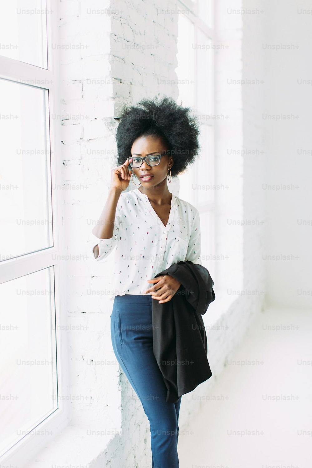 Young african american business woman in glasses, standing near the big window and holding jacket.