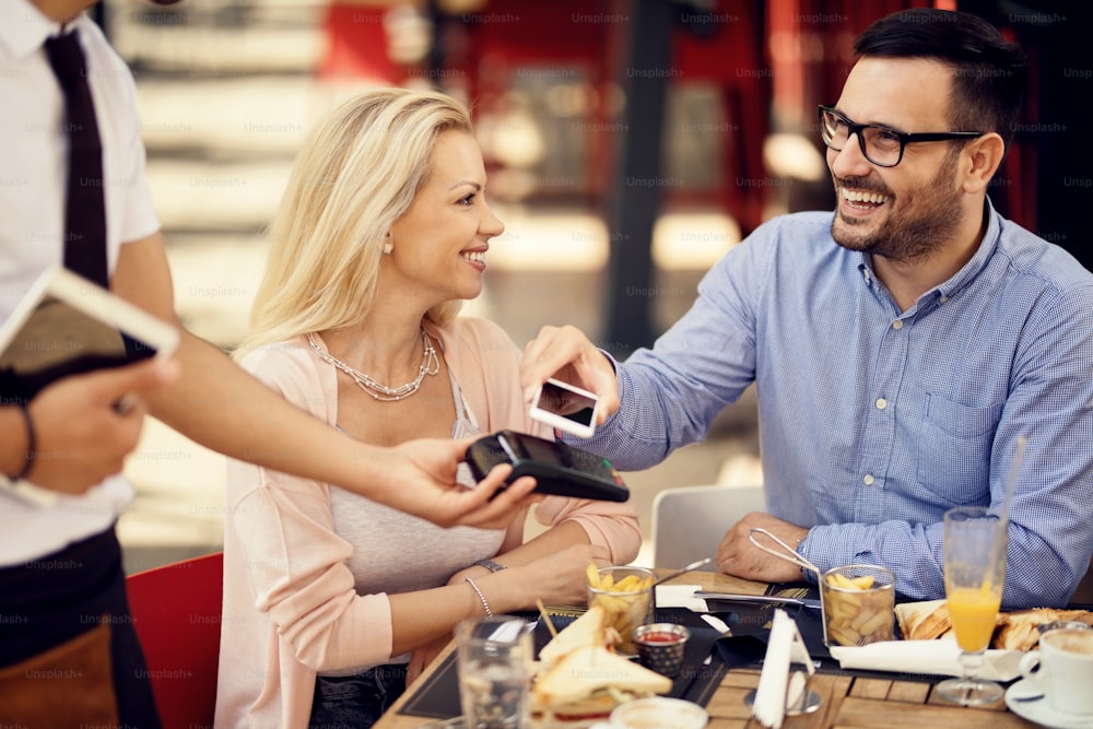 Uomo felice che posiziona lo smartphone sul lettore di carte di credito ed effettua il pagamento contactless in un bar.