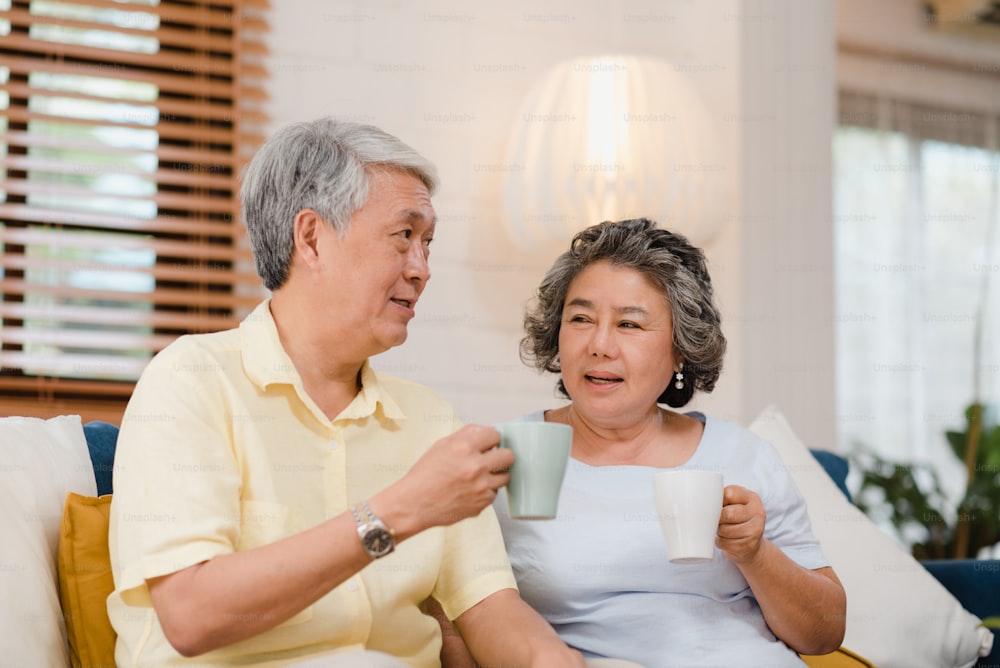 Pareja de ancianos asiáticos bebiendo café caliente y hablando juntos en la sala de estar de casa, la pareja disfruta del momento de amor mientras está acostada en el sofá cuando está relajada en casa. Estilo de vida de la familia mayor en el concepto de casa.