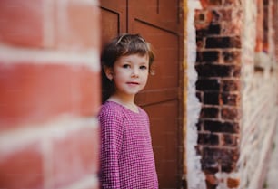 Un retrato de una pequeña niña linda parada al aire libre frente a la puerta, mirando a la cámara.