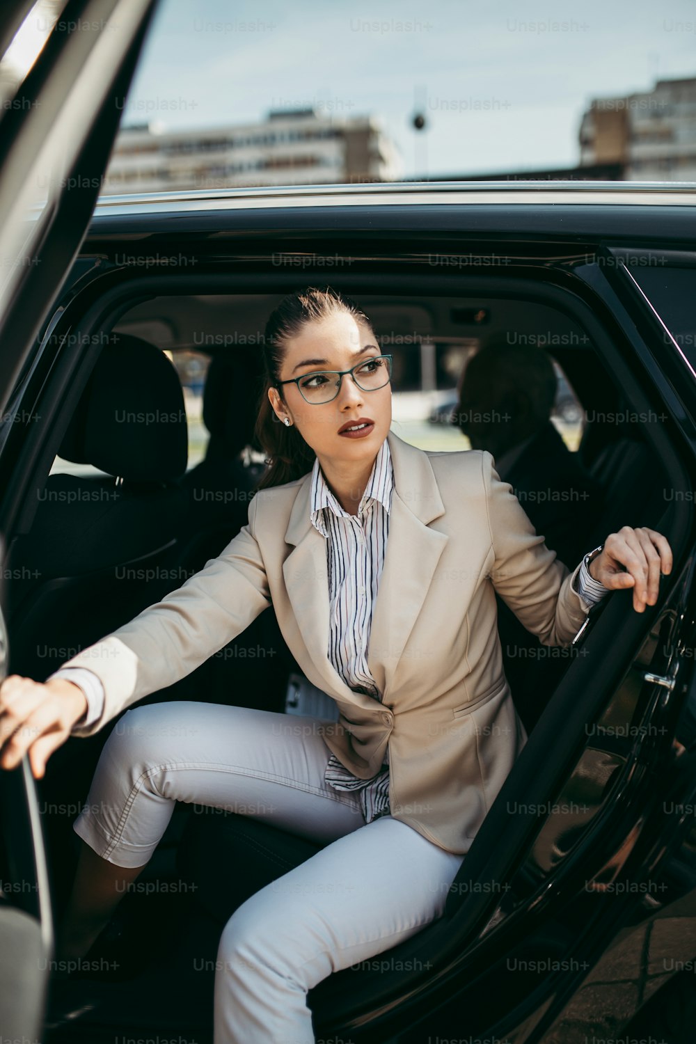 Gut aussehende junge Geschäftsfrau sitzt auf dem Rücksitz in Luxusauto. Sie benutzt ihr Smartphone, lächelt und schaut nach draußen. Transport im Unternehmenskonzept.
