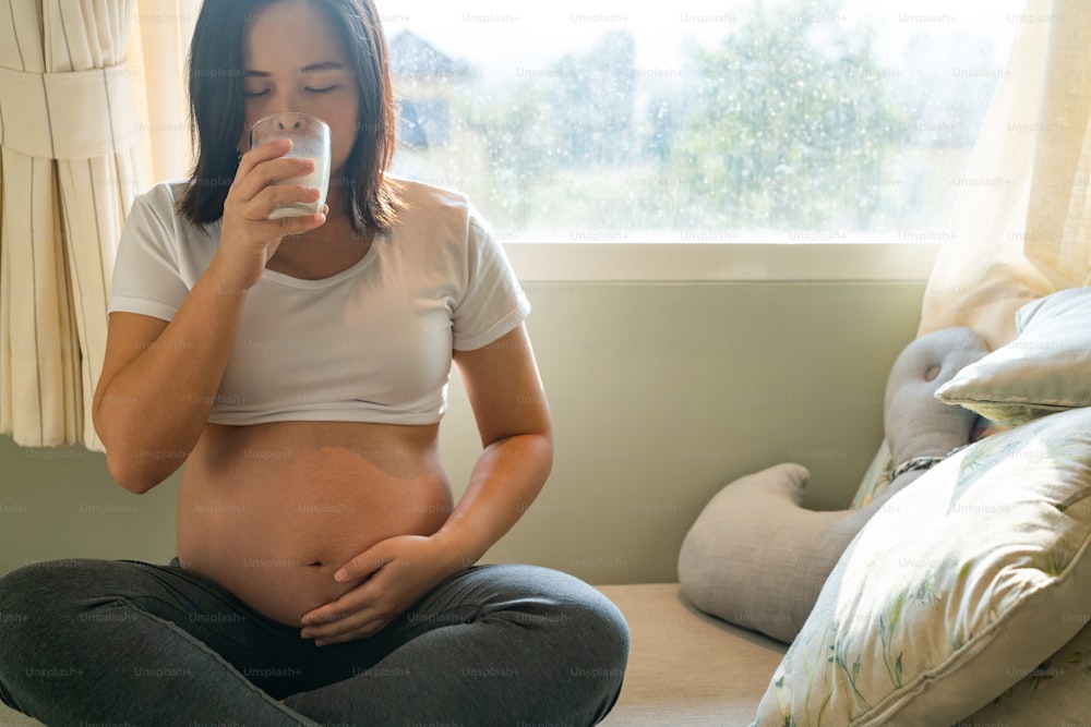 Happy pregnant woman drinks milk in glass at home while taking care of her child. The young expecting mother holding baby in pregnant belly. Calcium food nutrition for strong bones of pregnancy.