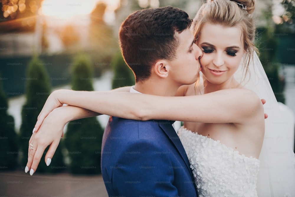 Stylish happy bride and groom kissing in warm sunset light at wedding reception outdoors. Gorgeous wedding couple of newlyweds  embracing in evening sunlight