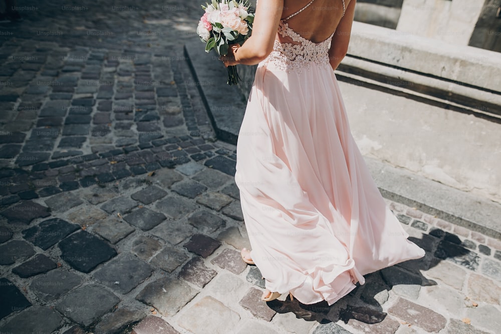 Stylish bridesmaid in pink dress walking in european city street and holding wedding bouquet, cropped view. Wedding celebration outdoors