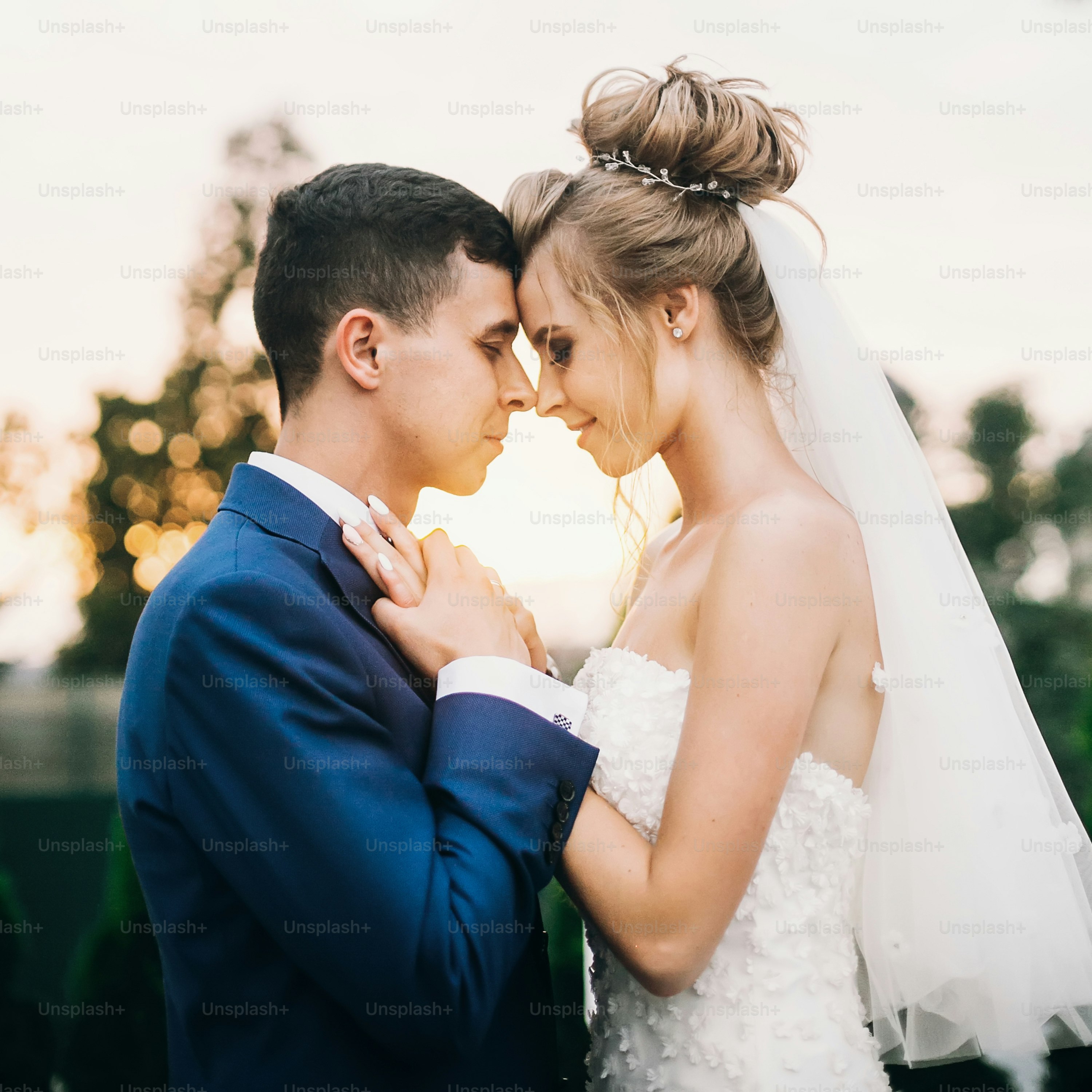 Stylish happy bride and groom posing in warm sunset light at wedding reception outdoors. Gorgeous wedding couple of newlyweds  embracing in evening sunlight