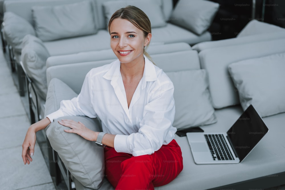 High angle of pretty smiling lady looking at camera while sitting near computer on sofa. Female model is dressing stylishly and posing for camera