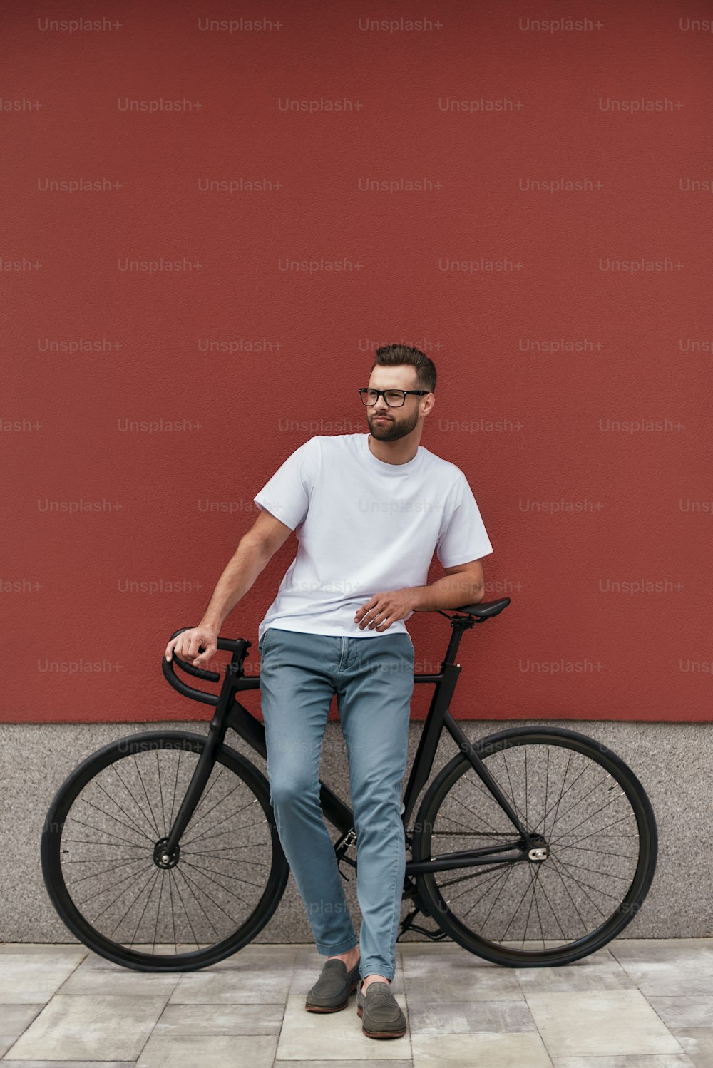 Active day. Handsome and young man with stubble in eyeglasses and casual clothes leaning at his bicycle and looking aside while standing against red wall outdoors. Weekend. City life