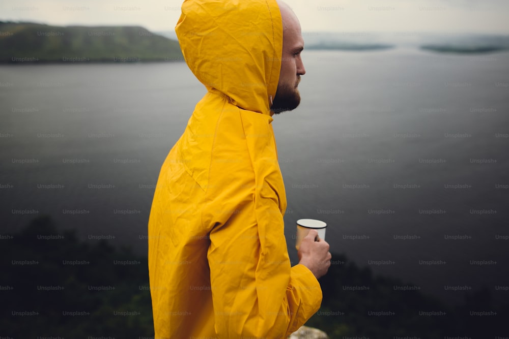 Traveler in yellow raincoat holding metal mug and standing on cliff  in rainy windy day with view on lake. Wanderlust and travel. Hipster man hiking in Norway. Atmospheric moment