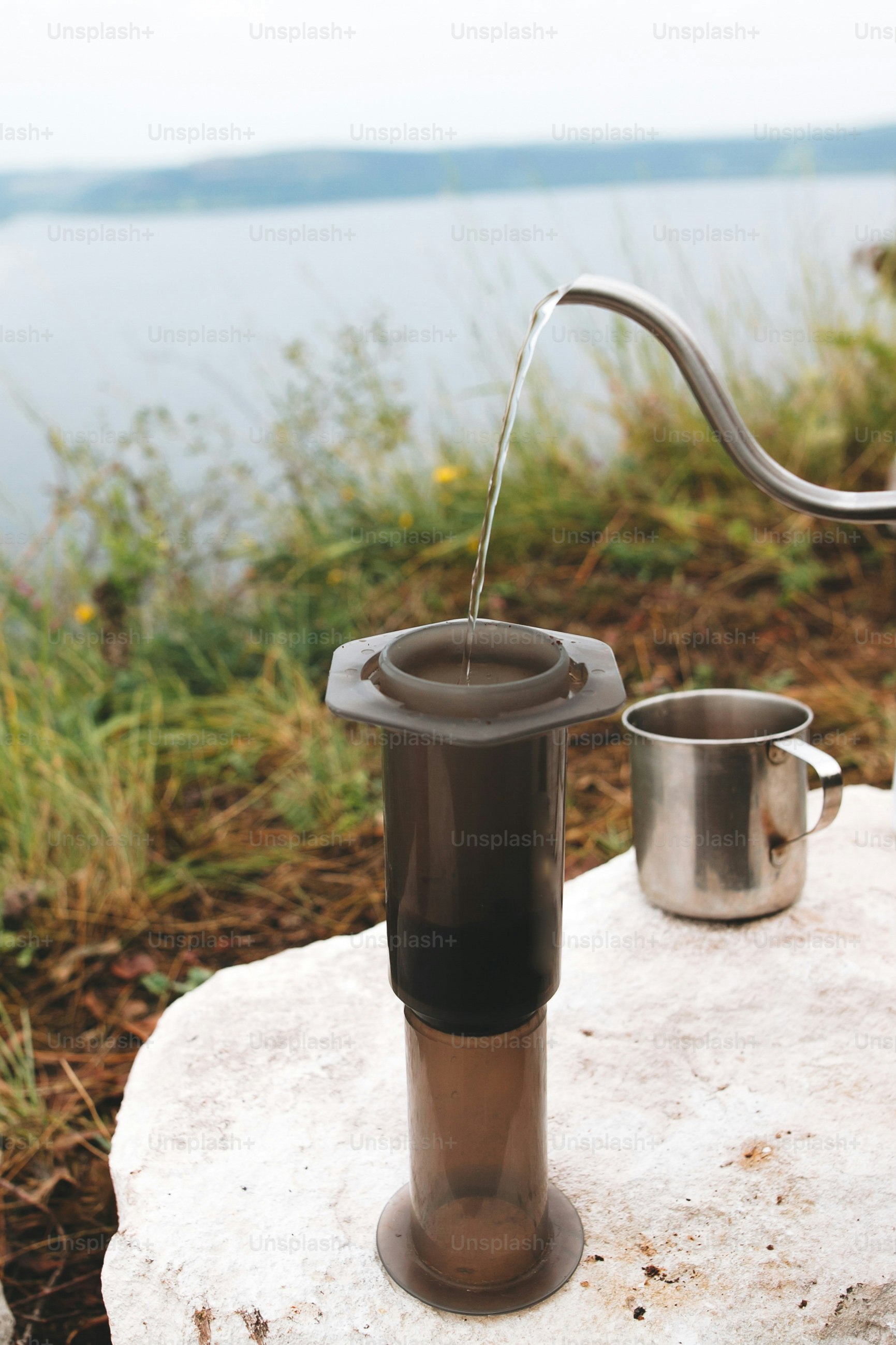 Brewing alternative coffee at camping. Traveler pouring hot water from steel kettle in aeropress on cliff at lake. Making hot drink at picnic outdoors. Trekking and hiking in mountains
