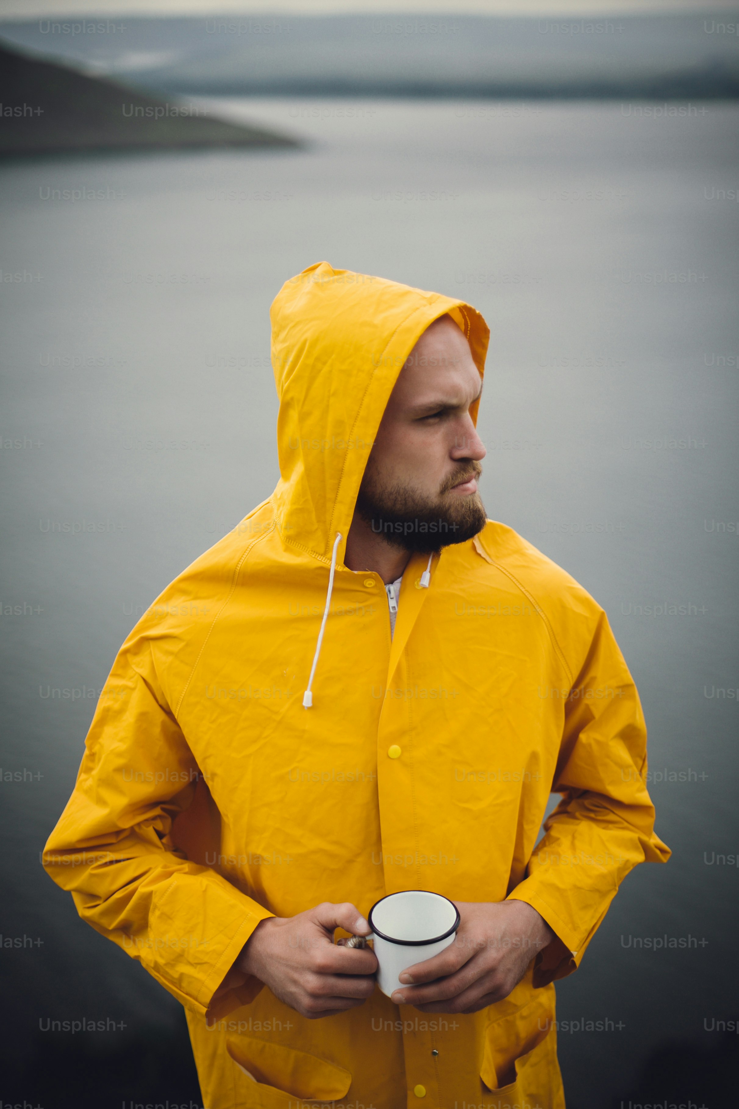 Wanderlust. Viajero hipster con impermeable amarillo sosteniendo una taza de metal y de pie en un acantilado en un día lluvioso y ventoso con vista al lago. Hombre hipster haciendo senderismo en Noruega. Momento atmosférico