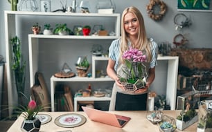 Female florist in flower shop standing with orchid in hands. Young beautiful blonde selling flowers and succulents.