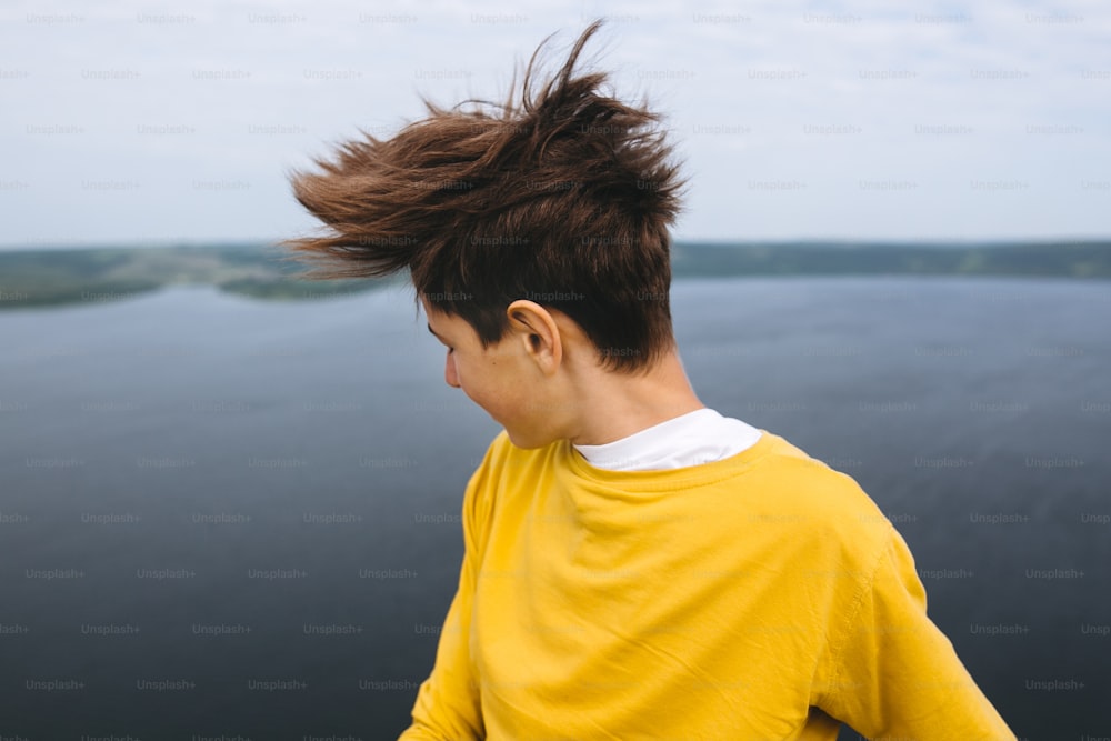 Stylish hipster teenager having fun and waving hair on top of rock mountain with amazing view on river. Young guy in yellow pullover exploring and traveling. Copy space