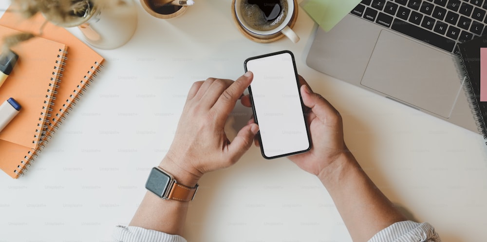 Top view of man holding blank screen smartphone in minimal workplace with laptop on white wooden table