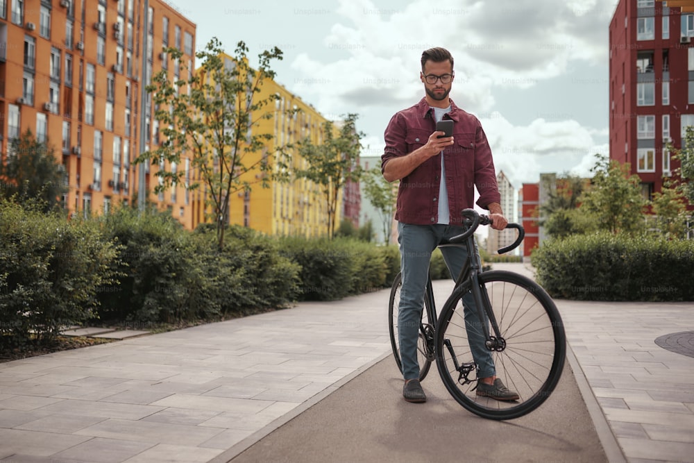 Chatting with friend. Full length of handsome man with stubble in casual clothes and eyeglasses holding mobile phone while walking with his bicycle outdoors. Digital concept. Weekend activity