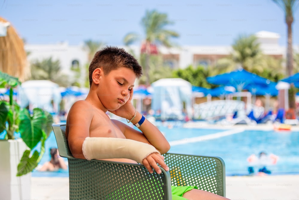 Niño triste con el brazo enyesado neer piscina, niño con brazo roto no puede disfrutar de sus vacaciones. Niño que no puede nadar en el agua con el brazo enyesado
