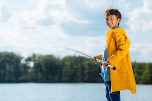 Fishing time. Handsome dark-haired boy wearing yellow rain coat smiling while fishing