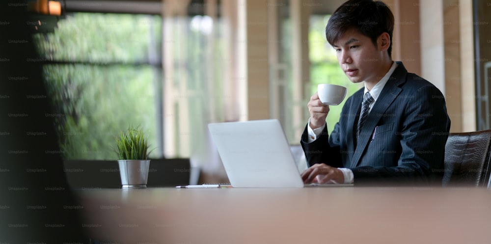 Jovem empresário atraente tendo uma xícara de café enquanto digita no laptop