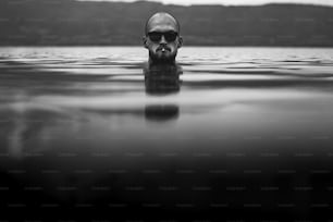 담배를 피우고 선글라스를 낀 잔인한 수염을 기른 남자가 호수에 나타납니다. 비오는 안개가 낀 날, 대기 순간에 호수에서 물 위에서 담배를 피우는 남자 머리. 방랑벽. 창조적 인 흑백 사진