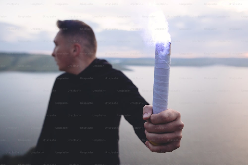 Brutal man in black windbreaker holding blue flare torch in hand, standing on top of rock in evening with view on river.  Ultras hooligan with fire signal bomb. Atmospheric moment