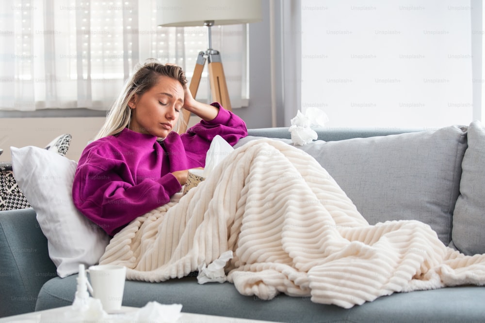 Sick woman with headache sitting under the blanket. Sick woman with seasonal infections, flu, allergy lying in bed. Sick woman covered with a blanket lying in bed with high fever and a flu, resting.