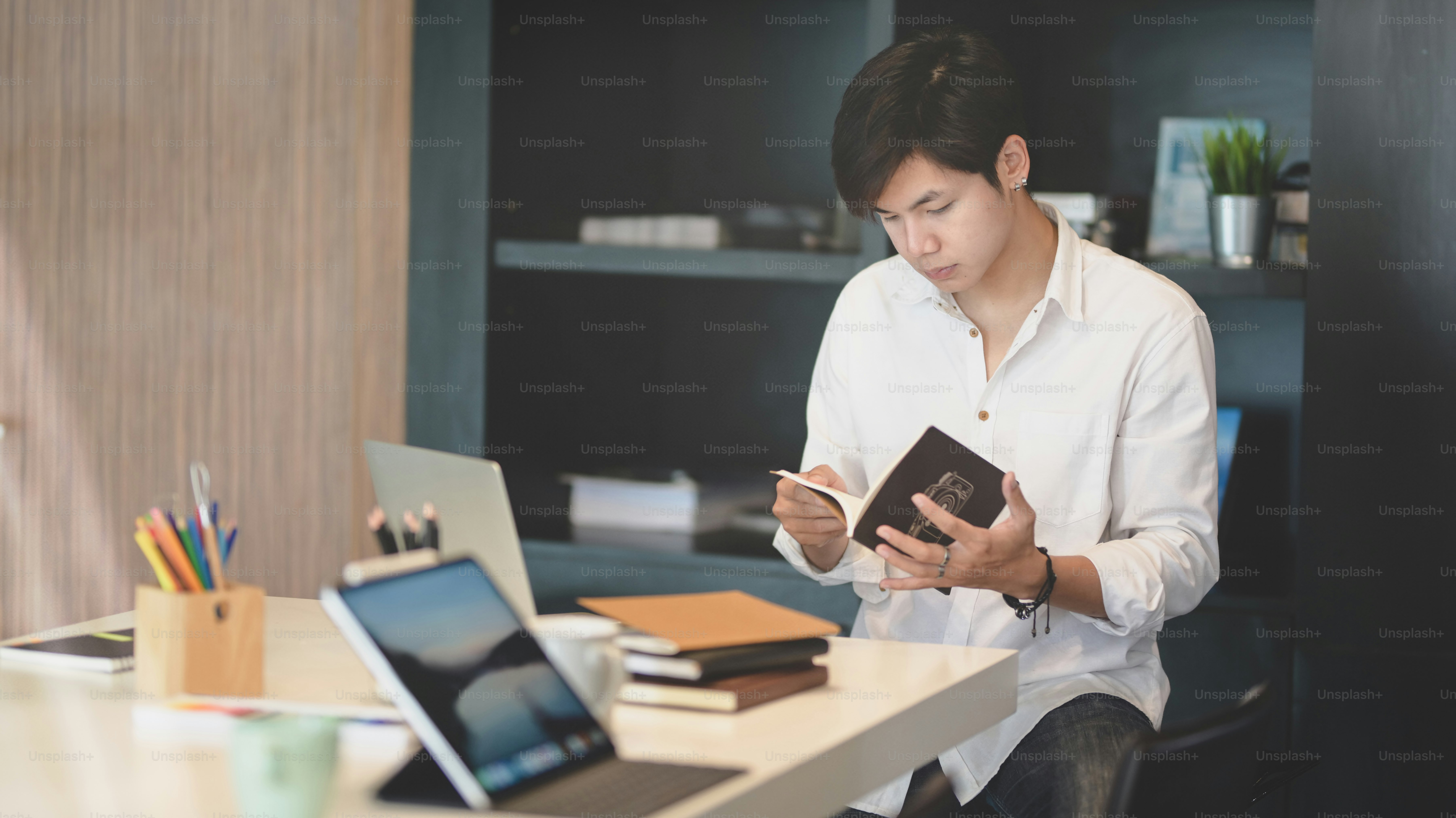 Professional young male freelancer working on his project while looking for idea from books in trendy workplace