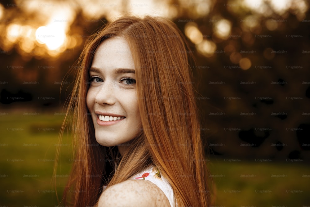 Close up portrait of a red hair woman girl with freckles looking at camera laughing over the shoulder against sunset outside.