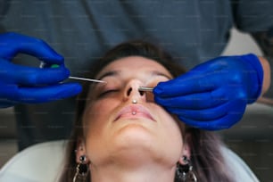 Portrait of a woman getting her nose pierced. Man showing a process of piercing nose with steril neadle and latex gloves. Nostril Piercing Procedure