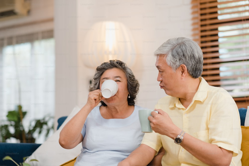 Pareja de ancianos asiáticos bebiendo café caliente y hablando juntos en la sala de estar de casa, la pareja disfruta del momento de amor mientras está acostada en el sofá cuando está relajada en casa. Estilo de vida de la familia mayor en el concepto de casa.