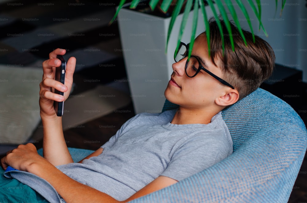 Concept de génération Z. Jeune garçon à lunettes utilisant un smartphone assis sur la chaise à l’intérieur.