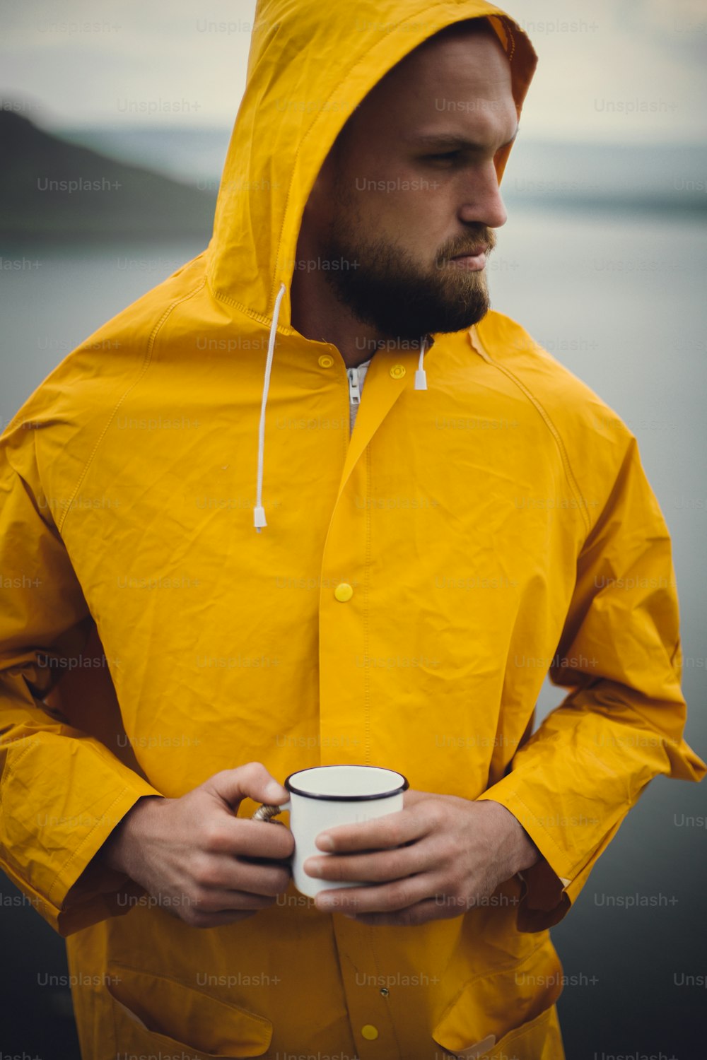 Wanderlust. Hipster traveler in yellow raincoat holding metal mug and standing on cliff  in rainy windy day with view on lake. Hipster man hiking in Norway. Atmospheric moment