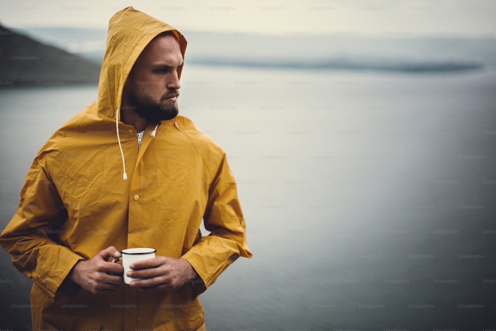 Atmospheric moment. Hipster traveler in yellow raincoat holding metal mug and standing on cliff in rainy windy day with view on lake. Wanderlust. Brutal bearded man hiking. Space