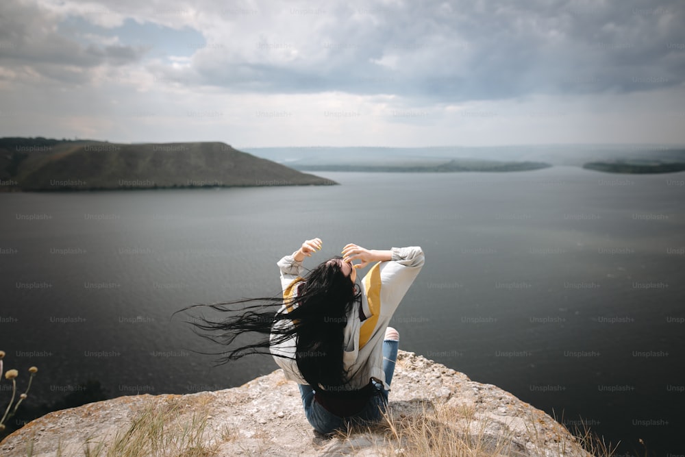 Espalda de una chica hipster con estilo sentada en la cima de la montaña de roca con una hermosa vista sobre el río. Joven turista con el pelo ventoso relajándose en el acantilado. Momento atmosférico. Espacio de copia
