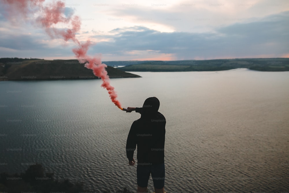 Ultras hooligan sosteniendo una bomba de humo rojo en la mano, de pie en la cima de la montaña rocosa con una increíble vista de la puesta de sol en el río. Momento atmosférico. Chico viajero con humo. Espacio de copia