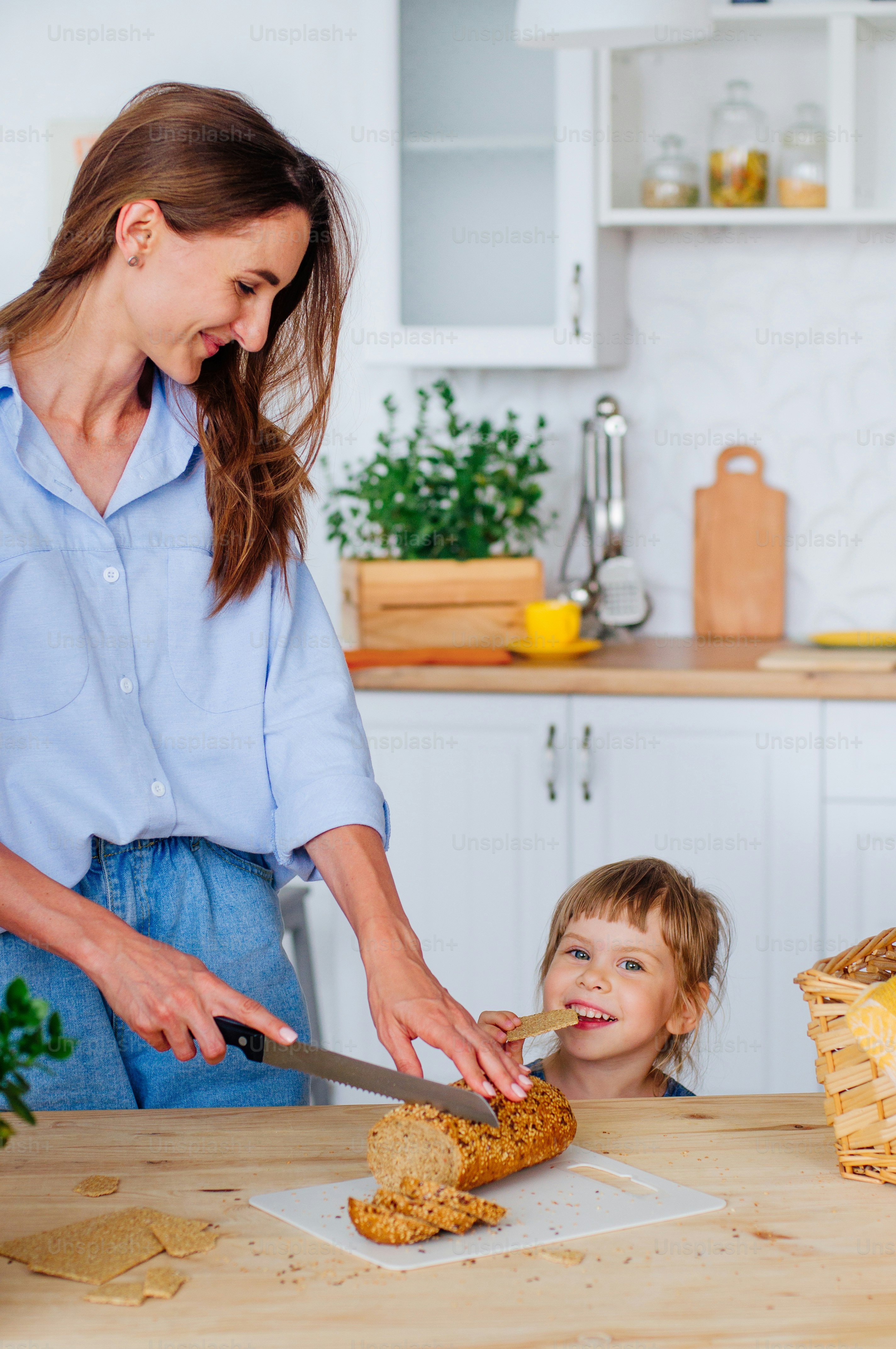 Happy little girl eating grain bread standing near her mother cutting bread at the table in the kitchen.