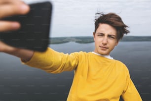 Stylish hipster teenager taking selfie on phone while standing on top of rock mountain with amazing view on river. Young guy in yellow pullover exploring and traveling. Copy space