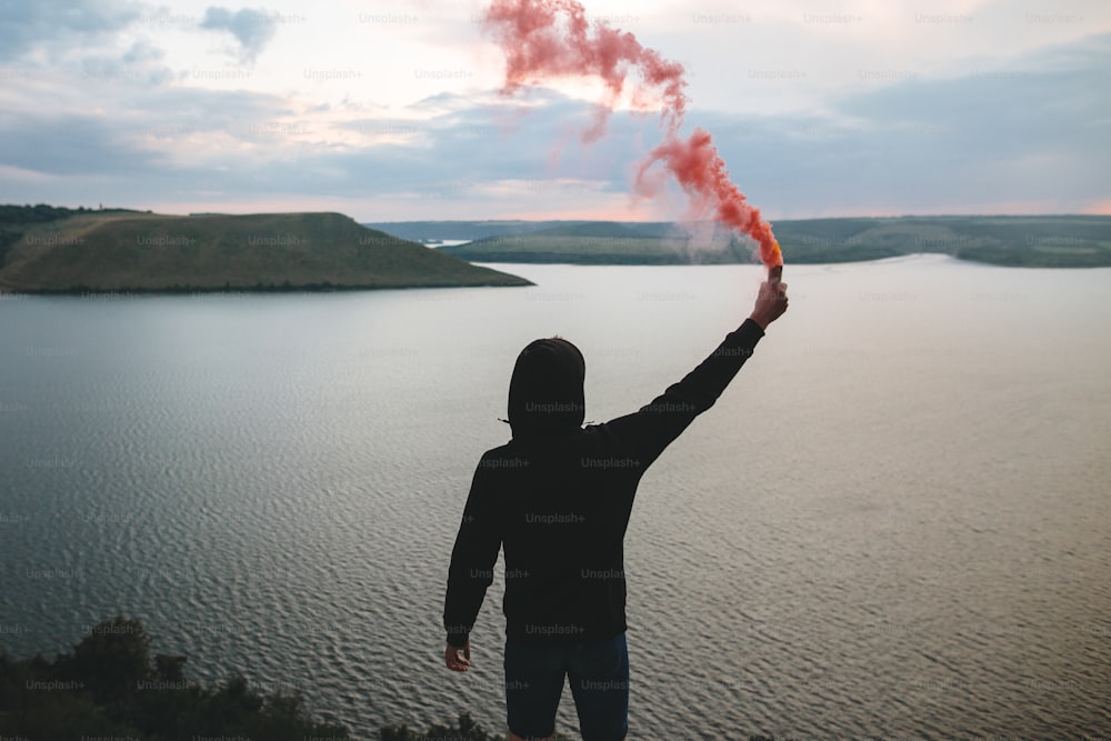Ultras hooligan holding red smoke bomb in hand, standing on top of rock mountain with amazing sunset view on river. Atmospheric moment. Traveler guy with smoke. Copy space