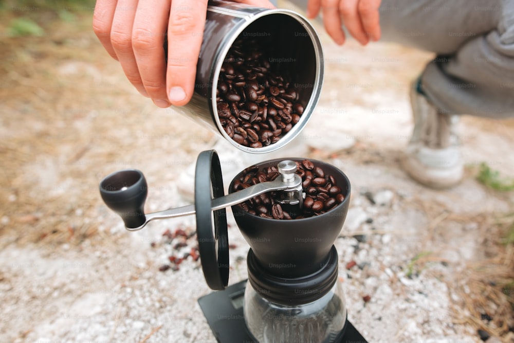 Filling grinder with fresh coffee beans on cliff at lake, preparing for brewing alternative coffee at camping. Making hot drink at picnic outdoors. Trekking and hiking in mountains