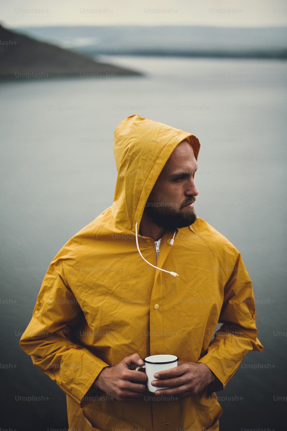 Brutal bearded man in yellow raincoat holding metal mug and traveling on cliff in rainy windy day with view on lake. Atmospheric moment. Wanderlust. Hipster man hiking in Norway